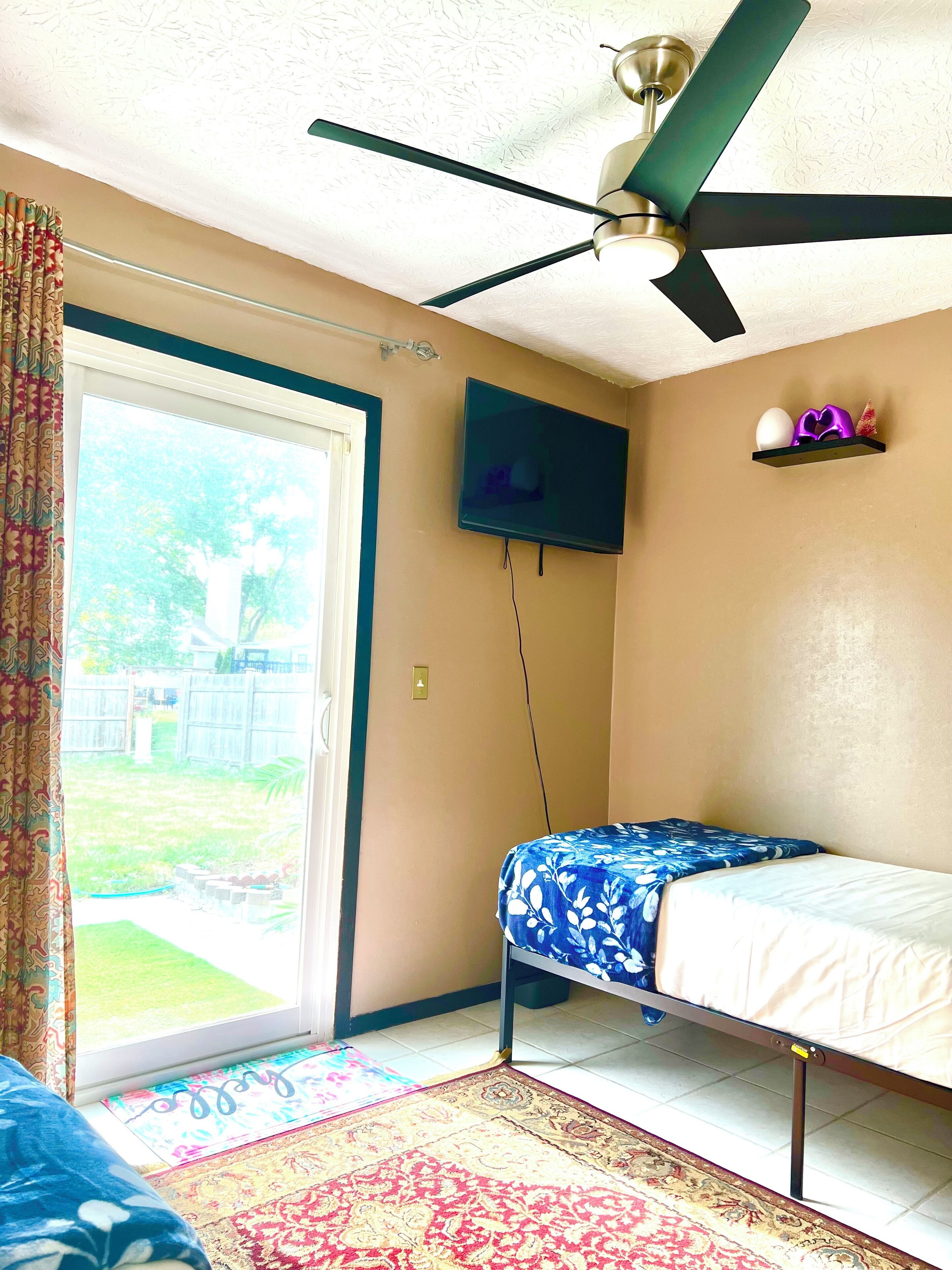 Una habitación con cama, ventilador de techo, televisor montado en la pared y vista al exterior.