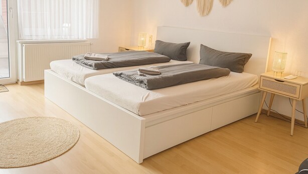 Una cama doble con cabecera blanca, dos mesitas de noche con lámparas, un tapete redondo tejido y una ventana con cortinas traslúcidas.