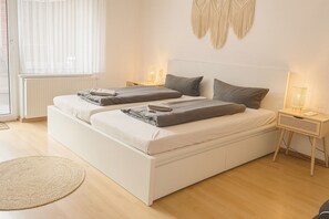 Un lit double avec une tête de lit blanche, deux tables de chevet avec des lampes, un tapis rond en fibres naturelles et une fenêtre avec des rideaux légers.
