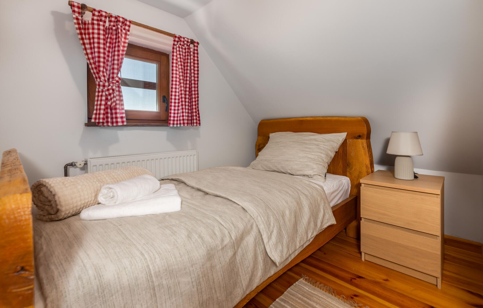 A bedroom with a wooden bed, a nightstand with a lamp, a window with checkered curtains, and a radiator.