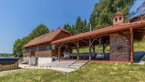A wooden cabin with a red-tiled roof, a covered patio with outdoor seating, and a stone wall. - Awesome home in Jaskovo with heated swimming pool (Jaskovo)