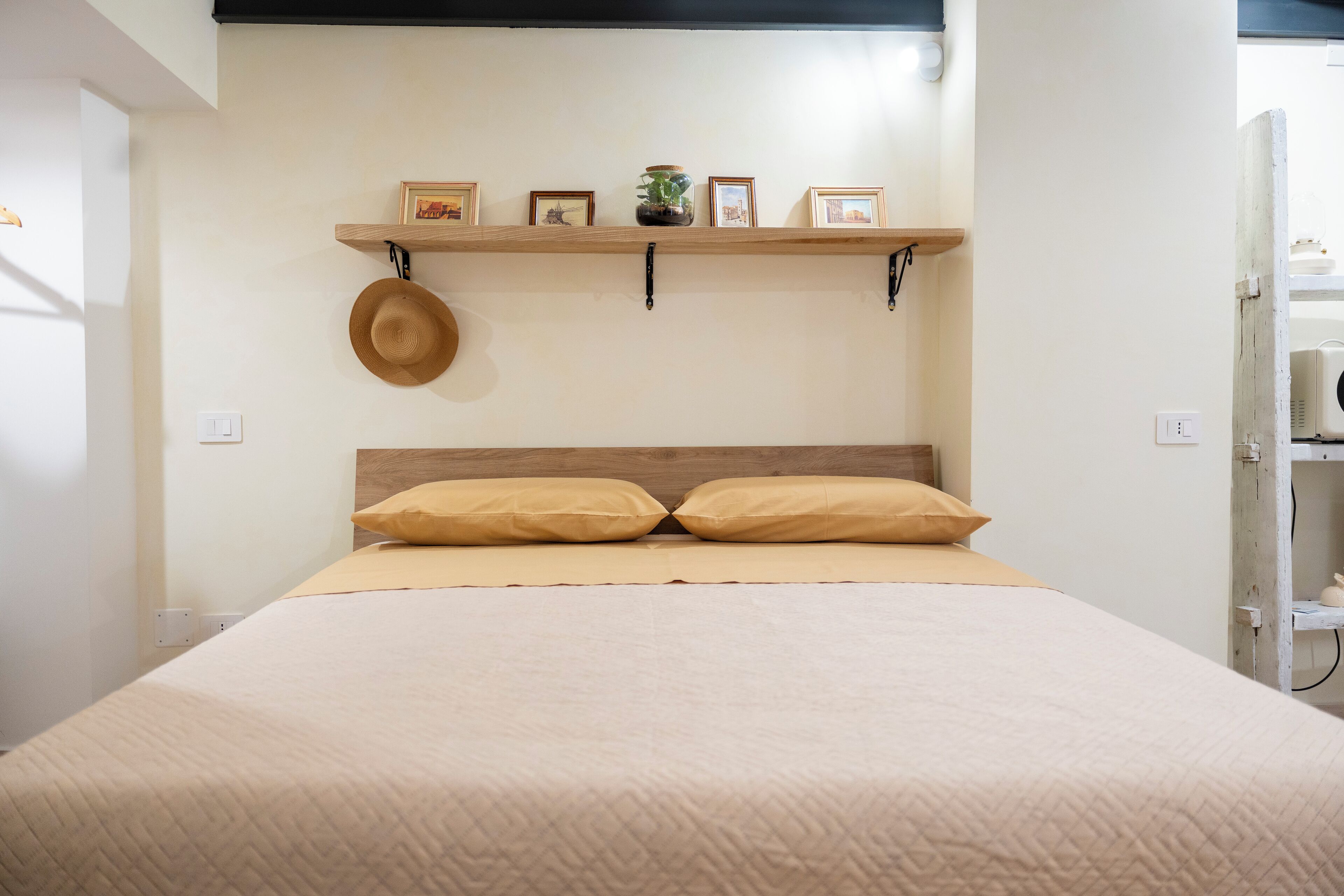 Een bed met een houten hoofdeinde, beige beddengoed en twee kussens. Daarboven een plank met ingelijste foto’s en een hoed.