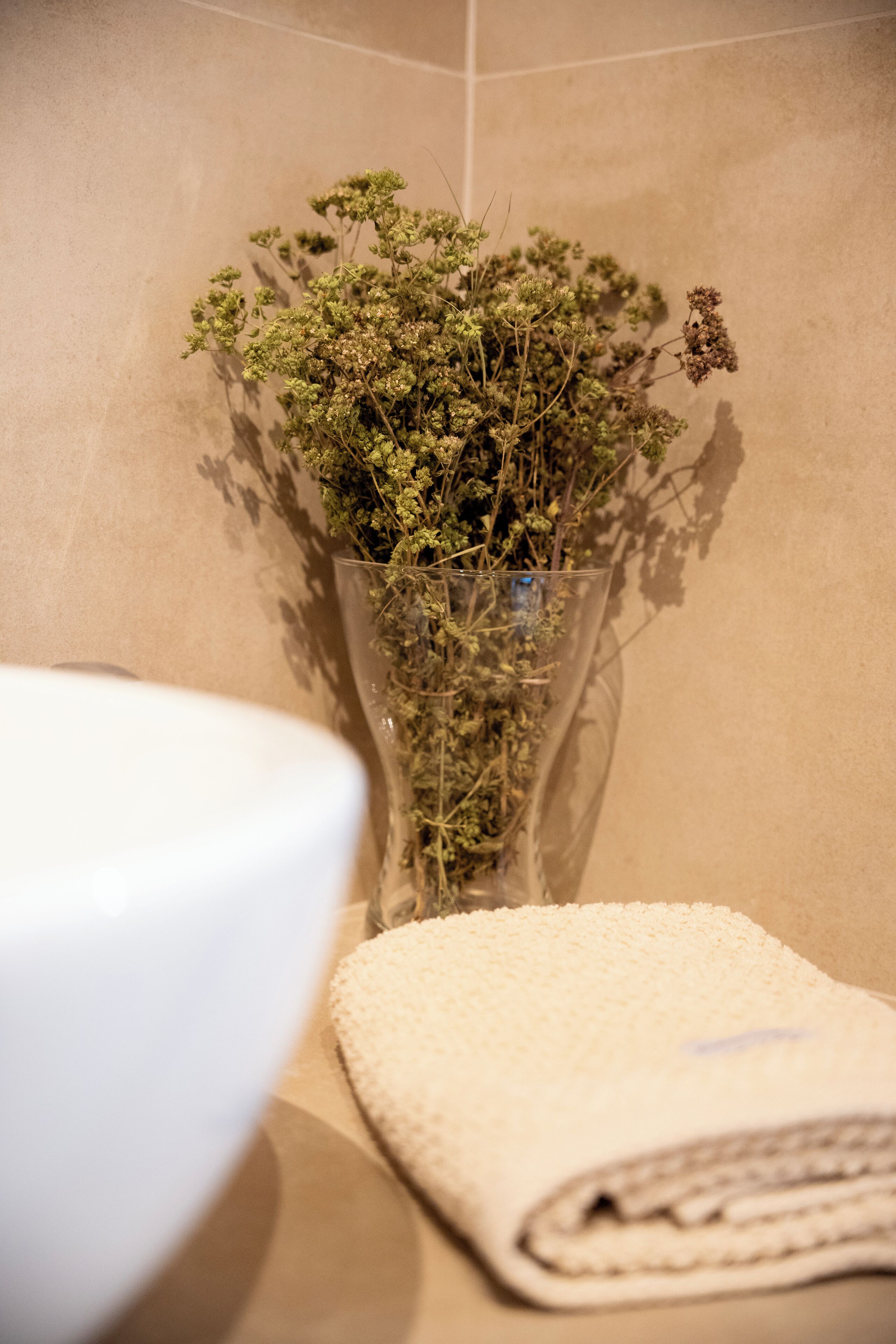 Une salle de bain avec un vase en verre contenant des fleurs séchées, posé sur un mur recouvert de carreaux beiges, un lavabo blanc et une serviette beige.