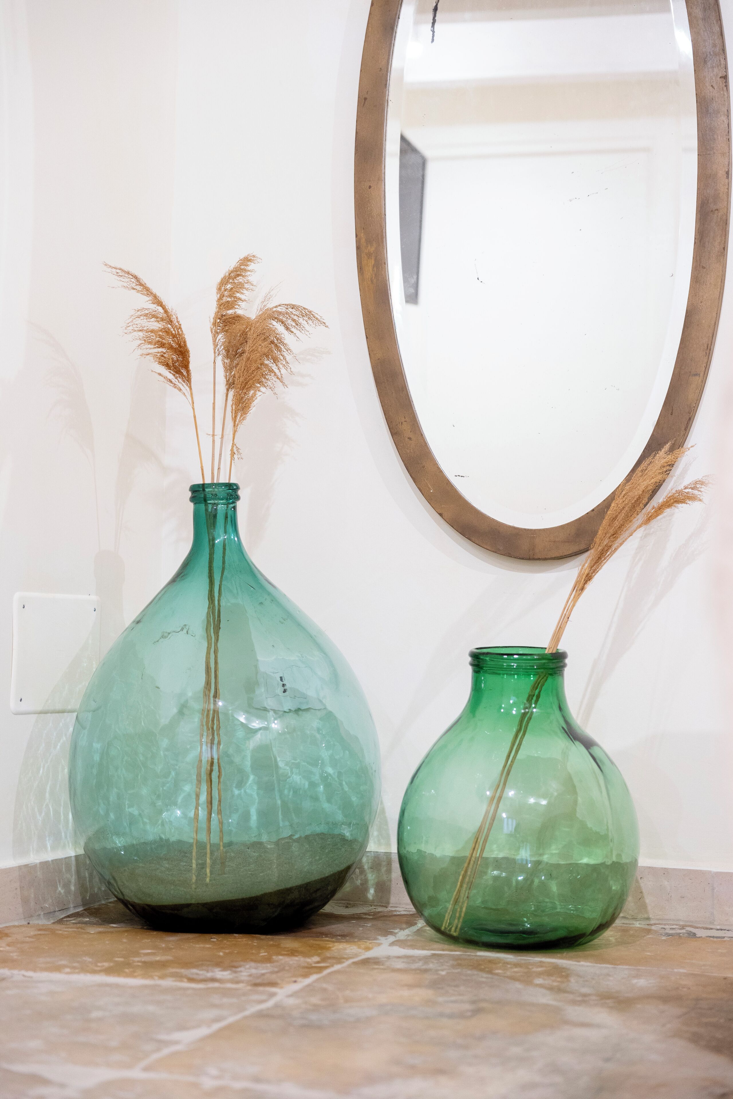 Two glass vases with dried plants on a tiled floor against a wall with a mirror.