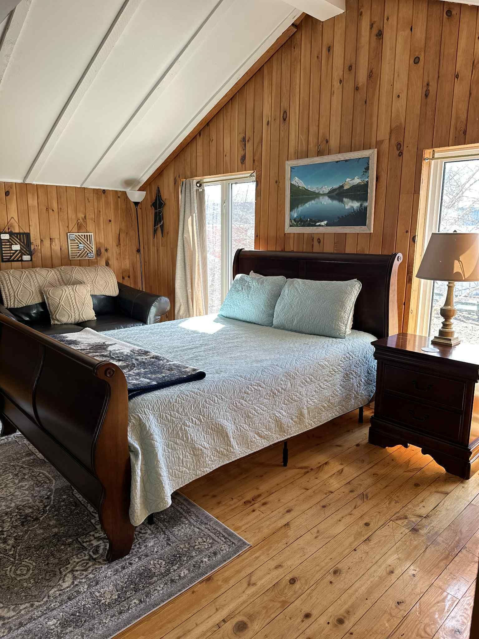 Ein gemütliches Schlafzimmer mit Holzboden, einem großen Bett mit Kopfteil, einem Nachttisch aus Holz und Seeblick durch ein Fenster.