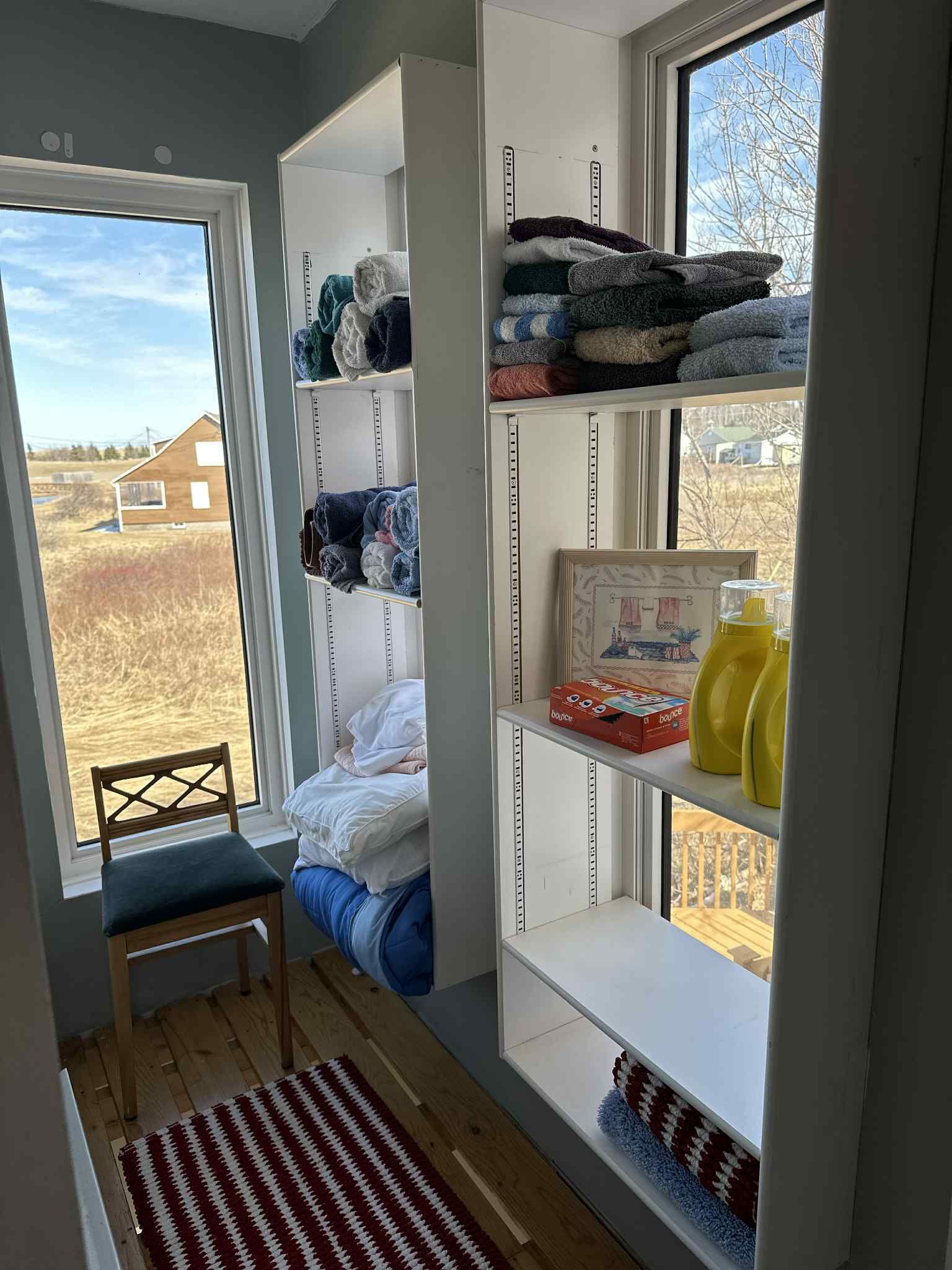 A room with a built-in shelving unit filled with folded towels, a small seating area with a chair, and a view of a rural landscape through a large window.