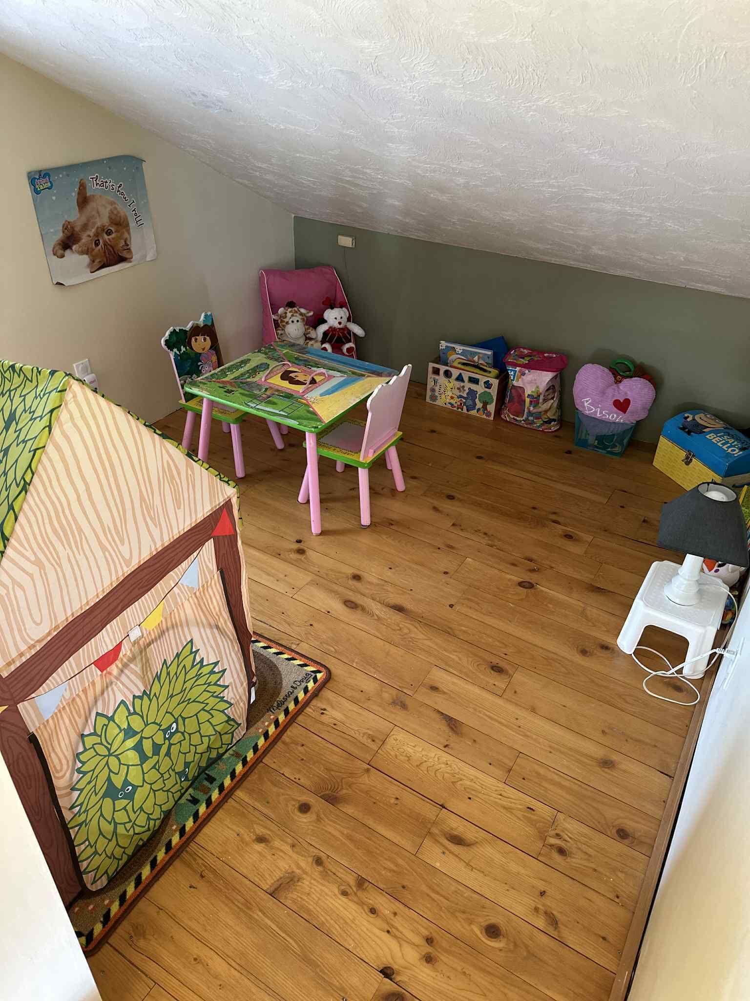 Une salle de jeux pour enfants, chaleureuse et accueillante, avec un plancher en bois, une petite maison de jeu, une chaise rose et divers jouets et boîtes de rangement.