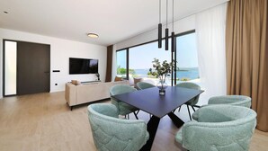 A modern living room with a large TV, a sofa, a dining table with chairs, and a view of the sea.