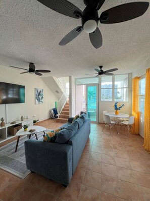 Ein geräumiges Wohnzimmer mit einem grauen Sofa, einem Fernseher und zwei Deckenventilatoren. Eine Treppe führt zu einem Außenbereich mit Blick auf Gebäude.
