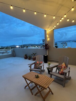 Une terrasse sur le toit, avec deux chaises en bois, une petite table, des guirlandes lumineuses, des plantes en pot et une vue sur le paysage urbain.