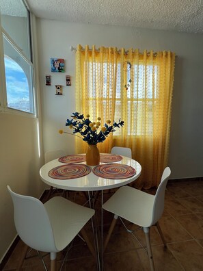 Un coin repas avec une table ronde, des chaises blanches, un vase avec des fleurs, des rideaux jaunes et de petits objets décoratifs accrochés au mur.