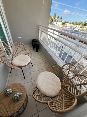 Ein Balkon mit Korbstühlen, einem kleinen Tisch, einer Brüstung und Blick auf Gebäude und Palmen.
