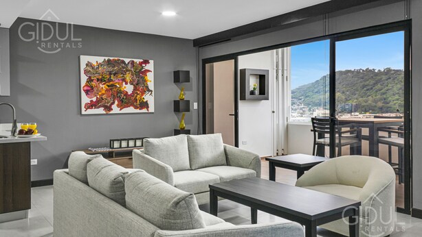 Una sala moderna con un sofá, una mesa de centro y un comedor con sillas y una mesa. Hay un ventanal con vistas a un paisaje urbano y colinas.
