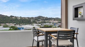 Un balcón con mesa de madera y cuatro sillas, con vistas a un paisaje urbano con edificios y áreas verdes.