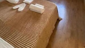 Ein Schlafzimmer mit Holzboden, einem Bett mit brauner Decke, einem Holzschrank und einem Fenster mit Vorhängen.