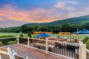 A deck with outdoor furniture, a barbecue grill, and string lights against a backdrop of a hilly landscape.
