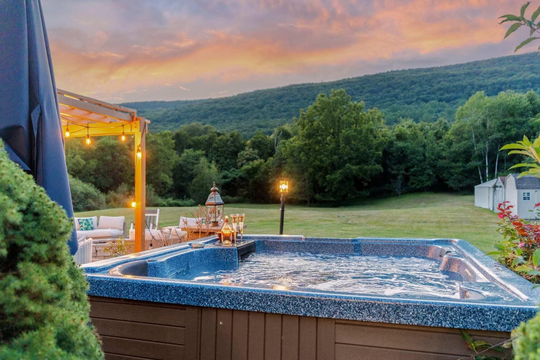 A hot tub with a pergola and string lights, surrounded by a lush green landscape and a house in the background.