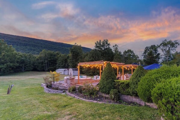 A gazebo with string lights, outdoor seating, and a view of a forested hill.