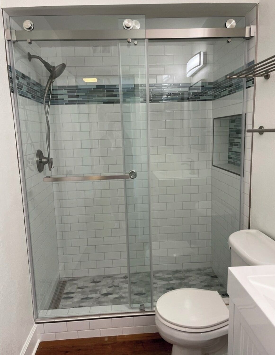 Ducha con cerramiento de vidrio, pared con azulejos, marco de puerta de madera y un inodoro blanco.