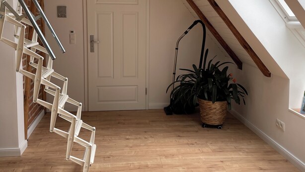 A spacious loft with a sloped ceiling, a wooden staircase, and a large window providing natural light.