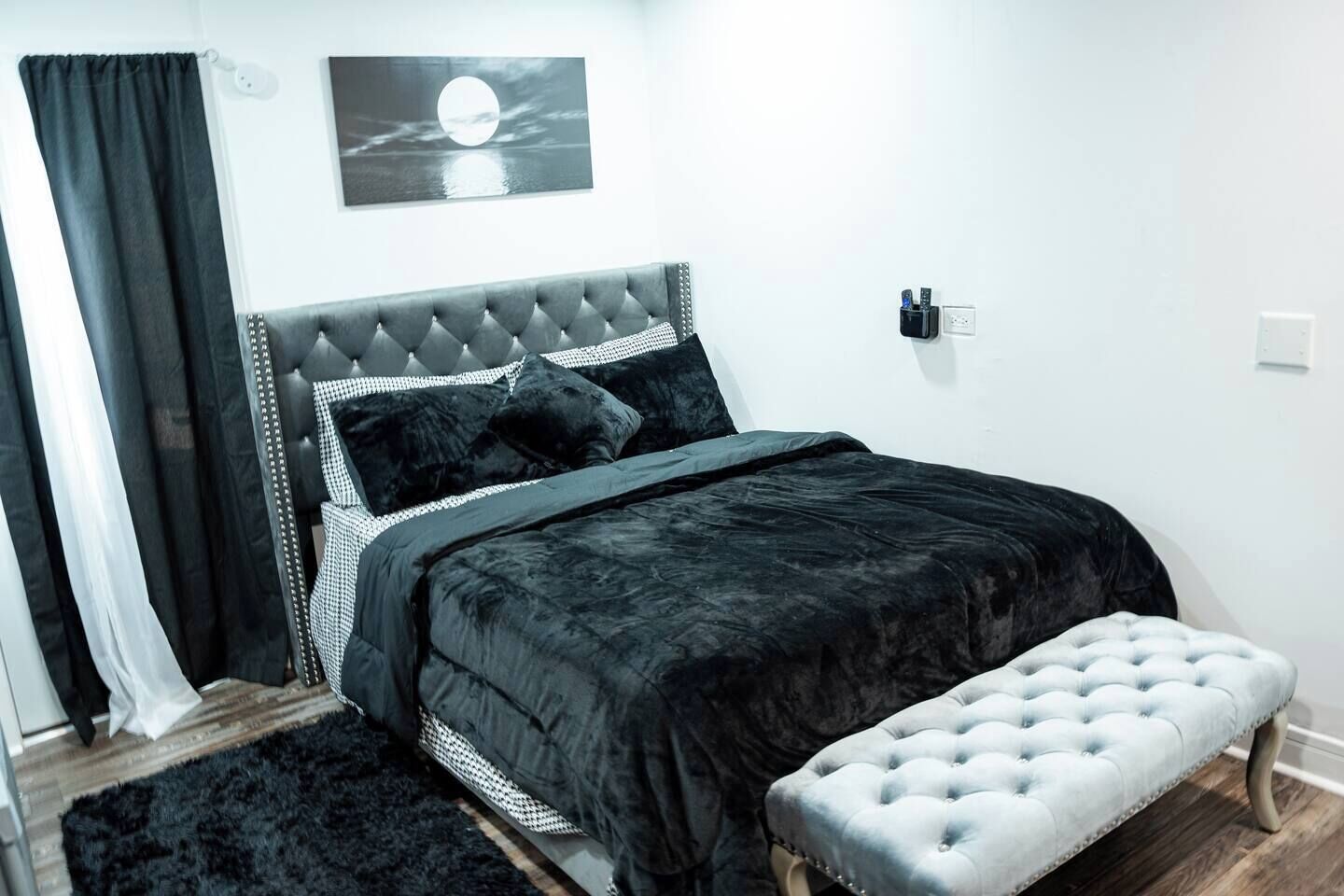 Quarto com cabeceira estofada, uma cama grande com roupa de cama escura, um banquinho e um candeeiro fixado na parede.