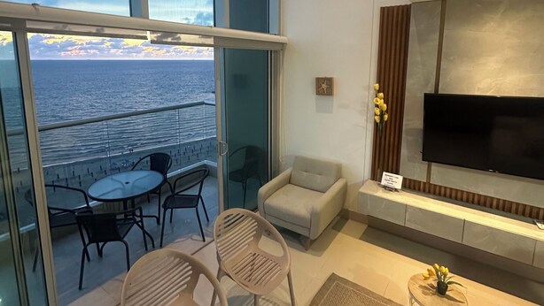 Ein modernes Wohnzimmer mit großem Fernseher, bequemen Sitzgelegenheiten, einem Glastisch und Blick auf das Meer.