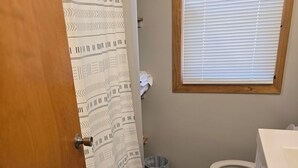 Une salle de bain comprenant des toilettes, un lavabo, une fenêtre avec des stores, un rideau de douche à motifs et un tapis de sol à motifs.