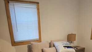Un lit bien fait, avec une couette et des oreillers de couleur beige, une table de chevet avec une lampe et une fenêtre équipée de stores.