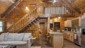 Un vaste chalet de style loft, doté d’un intérieur en bois, d’un escalier menant à un niveau supérieur, d’un coin cuisine et d’un espace salon avec un canapé et une table basse.