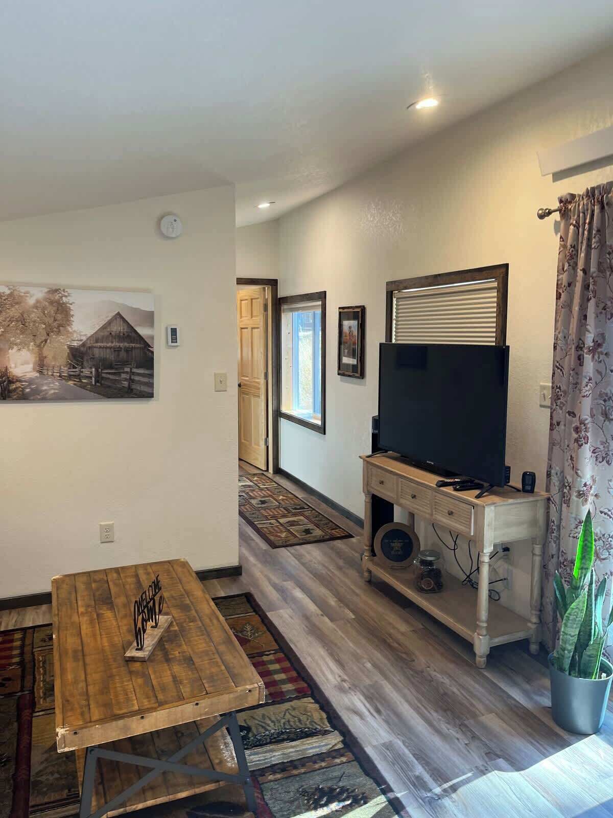 Un salón con una mesa de madera, un televisor sobre un mueble bajo y un cuadro en la pared.