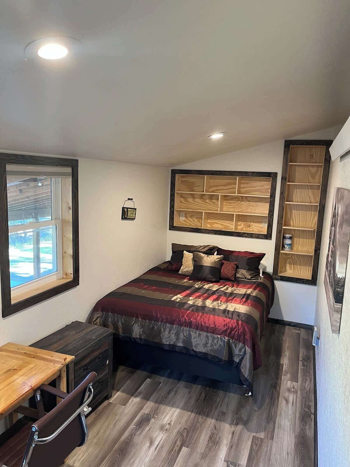 A bedroom with a bed, a wooden desk, a chair, a window, and a built-in wooden shelf.