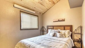 Una camera da letto con soffitto in legno, un letto con copripiumino decorato, una finestra con persiane e una mensola a muro con oggetti decorativi.