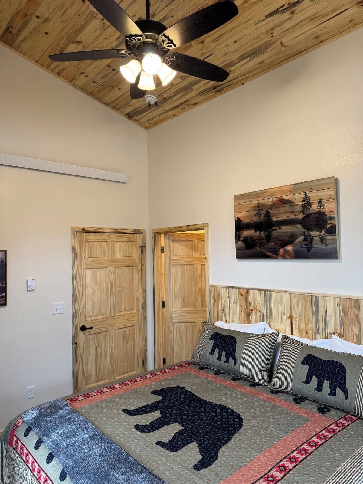 A bedroom with a wooden ceiling, a ceiling fan, a bed with a bear-themed bedspread, two wooden doors, and a landscape painting on the wall.