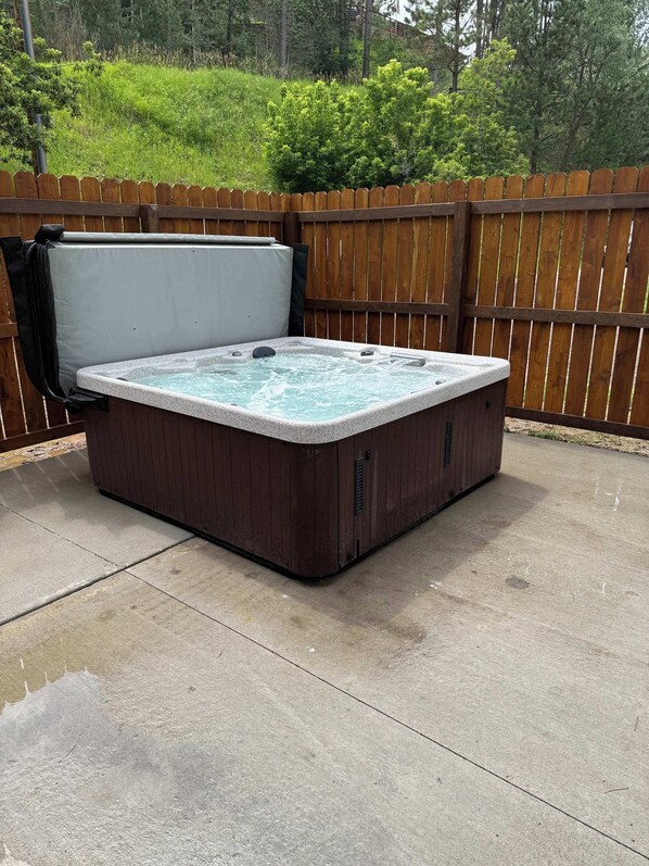 A hot tub with a wooden frame and a concrete base, surrounded by a wooden fence and greenery.