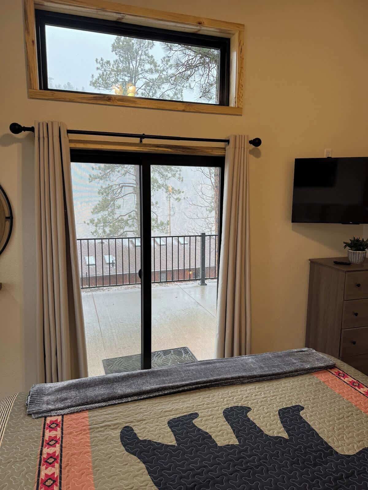 A room with a large window, a sliding door leading to an outdoor area, a bed with a patterned bedspread, a television on the wall, and a wooden cabinet.
