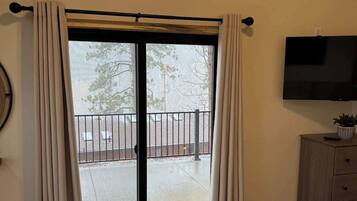 A room with a large window, a sliding door leading to an outdoor area, a bed with a patterned bedspread, a television on the wall, and a wooden cabinet.