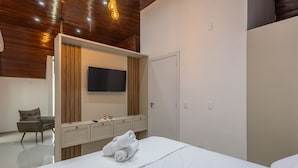 Una habitación de hotel moderna con techo de madera, un televisor de pantalla plana montado en la pared, un minibar, una cama con una toalla enrollada y una silla.