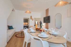 A modern dining area with a wooden table set for four, white chairs, and a television mounted on the wall. - La Douceur - Wifi - Proche gare (Laval)
