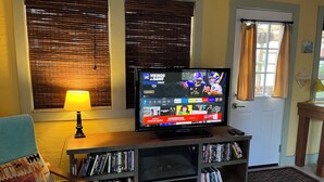 A living room with a flat-screen TV on a wooden stand, a bookshelf with DVDs, a lamp, and a door with curtains.