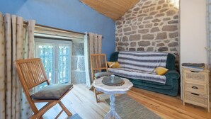 A cozy room with a stone wall, a wooden ceiling, a sofa with cushions, a wooden chair, a small table, and a window with curtains.