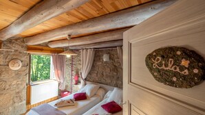 A cozy bedroom with two beds, stone walls, wooden beams, a stone fireplace, and a window with curtains.