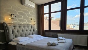 Een slaapkamer met een gestuukt hoofdeinde, een groot bed met witte lakens, uitzicht op de stad door een raam en een radiator op de vloer.