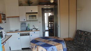 Cocina compacta con microondas, lavaplatos y fregadero. Una cama con manta estampada está en primer plano, y un sofá al fondo.