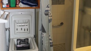 Un baño con ducha, lavamanos, estante con productos de limpieza y un cubo de basura.