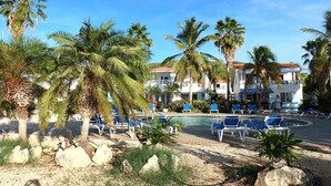 A pool area with palm trees, lounge chairs, and a building with a red roof in the background.