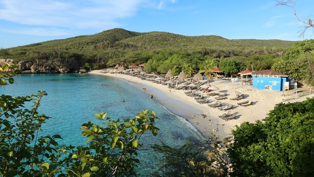 A beach with clear blue water, a sandy shore, and a few buildings in the background.