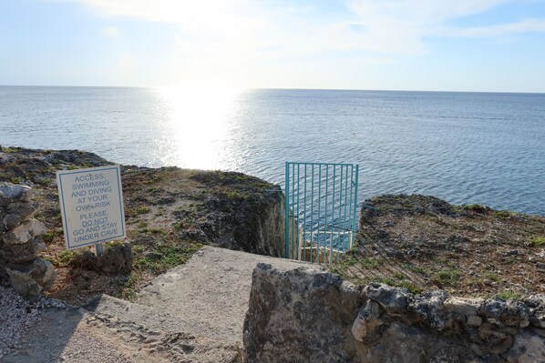 Un sendero costero con un cartel de advertencia sobre riesgos de natación y buceo, una reja metálica y una vista despejada del mar.