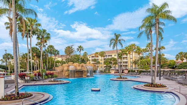 A large outdoor swimming pool with palm trees and a waterfall feature, surrounded by lounge chairs and a hotel building in the background. - 2-bedroom Resort Villa in Orlando-Universal, Disney, Epcot (Orlando)