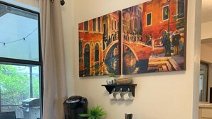 A room with a black console table, a coffee maker, a potted plant, and a wall-mounted shelf with cups.