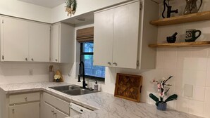 A kitchen with white cabinets, a marble countertop, a window with blinds, and a small shelf with kitchen tools.